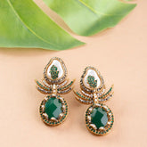 Ornate earrings with green gemstones on a beige surface with green leaves.