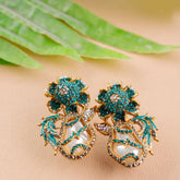 Ornate earrings with green gemstones and gold accents on a beige surface with green leaves.
