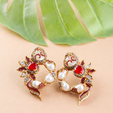 Pair of ornate earrings with red and white stones on a beige background with green leaves.