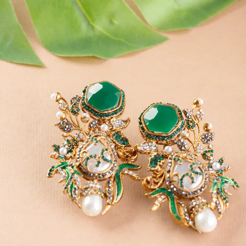 Ornate gold earrings, green stones, and pearls on a beige surface
