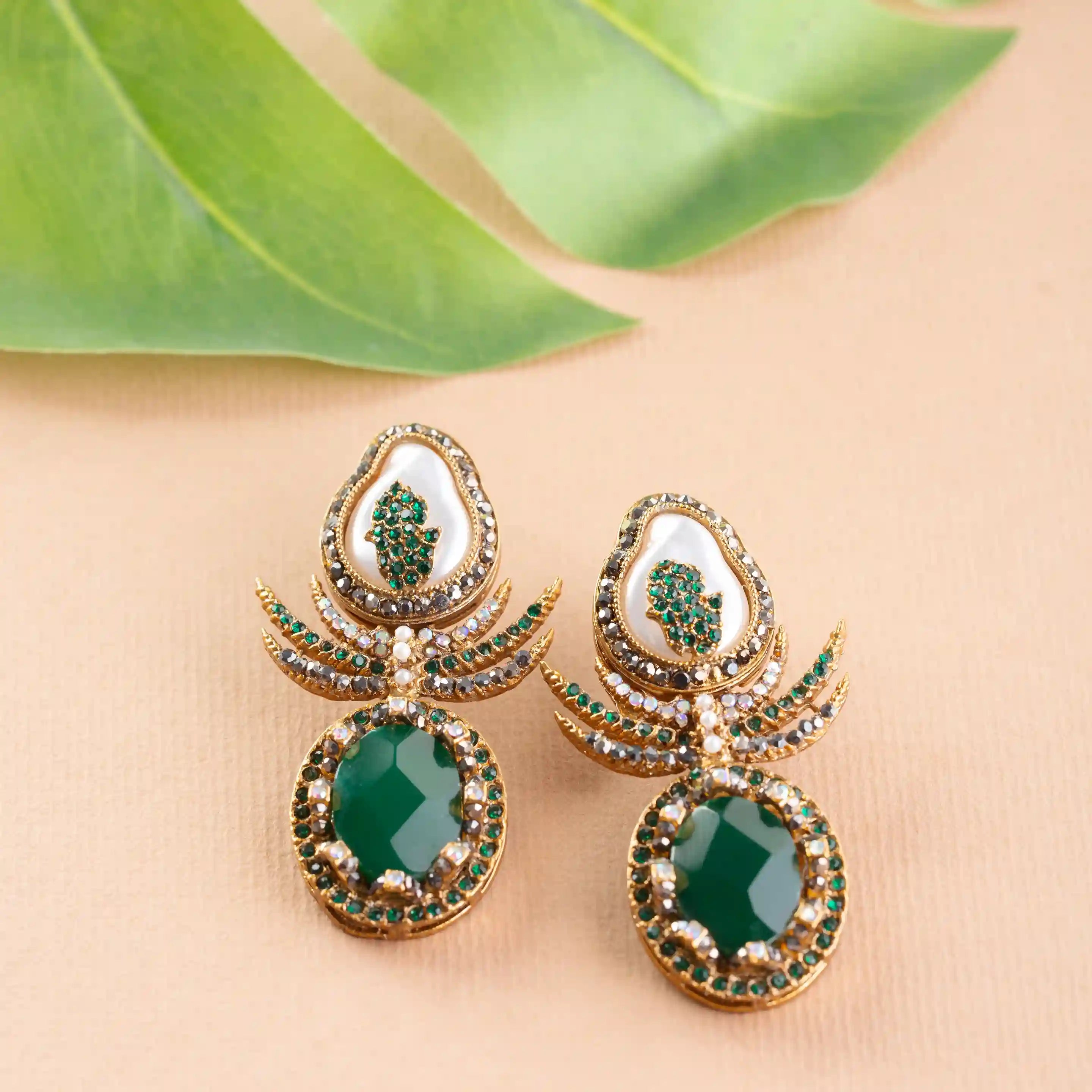 Ornate earrings with green gemstones on a beige surface with green leaves.