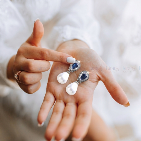 Person holding two pearl earrings with blue stones on a blurred background