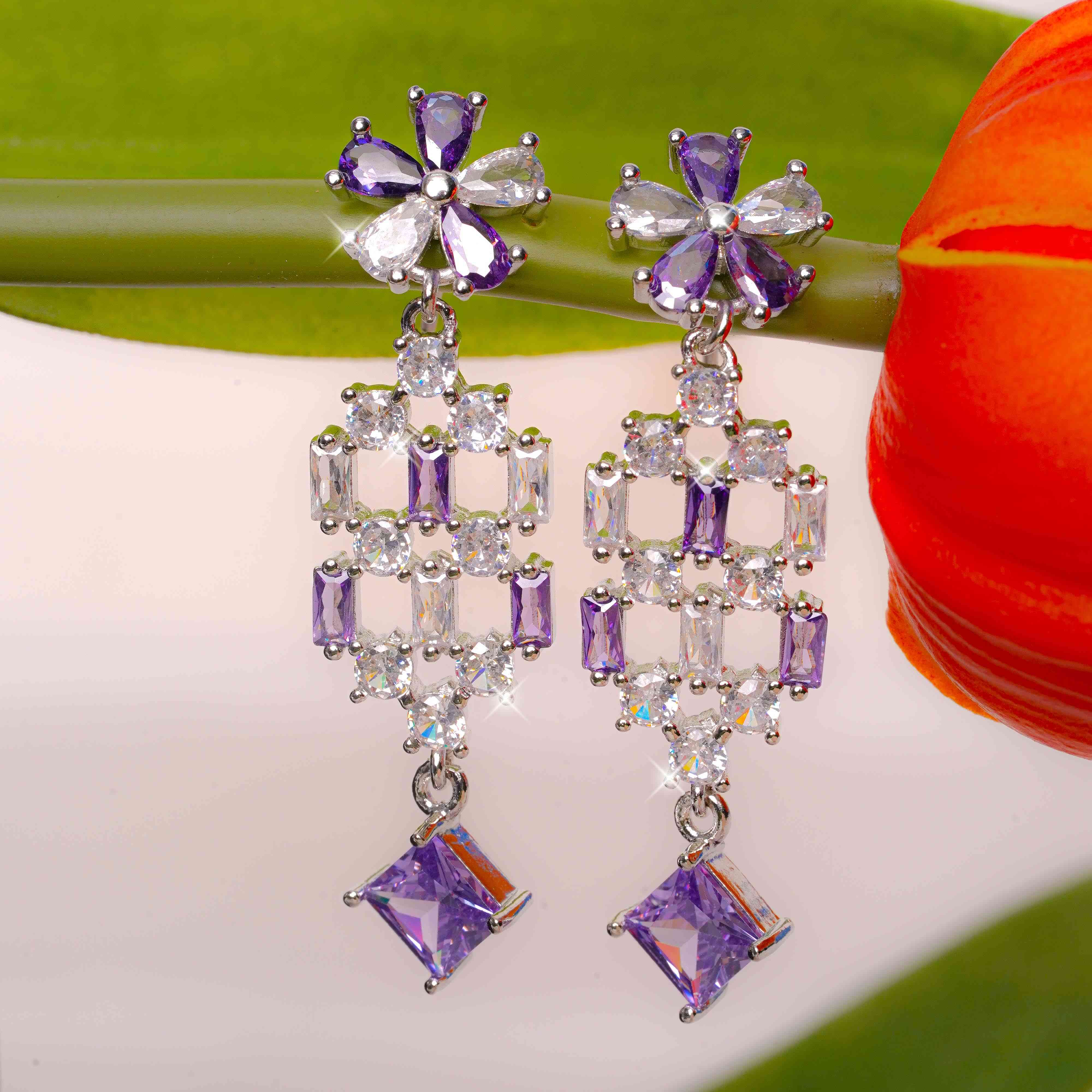 Pair of purple and clear gemstone earrings on a light background with a red flower.