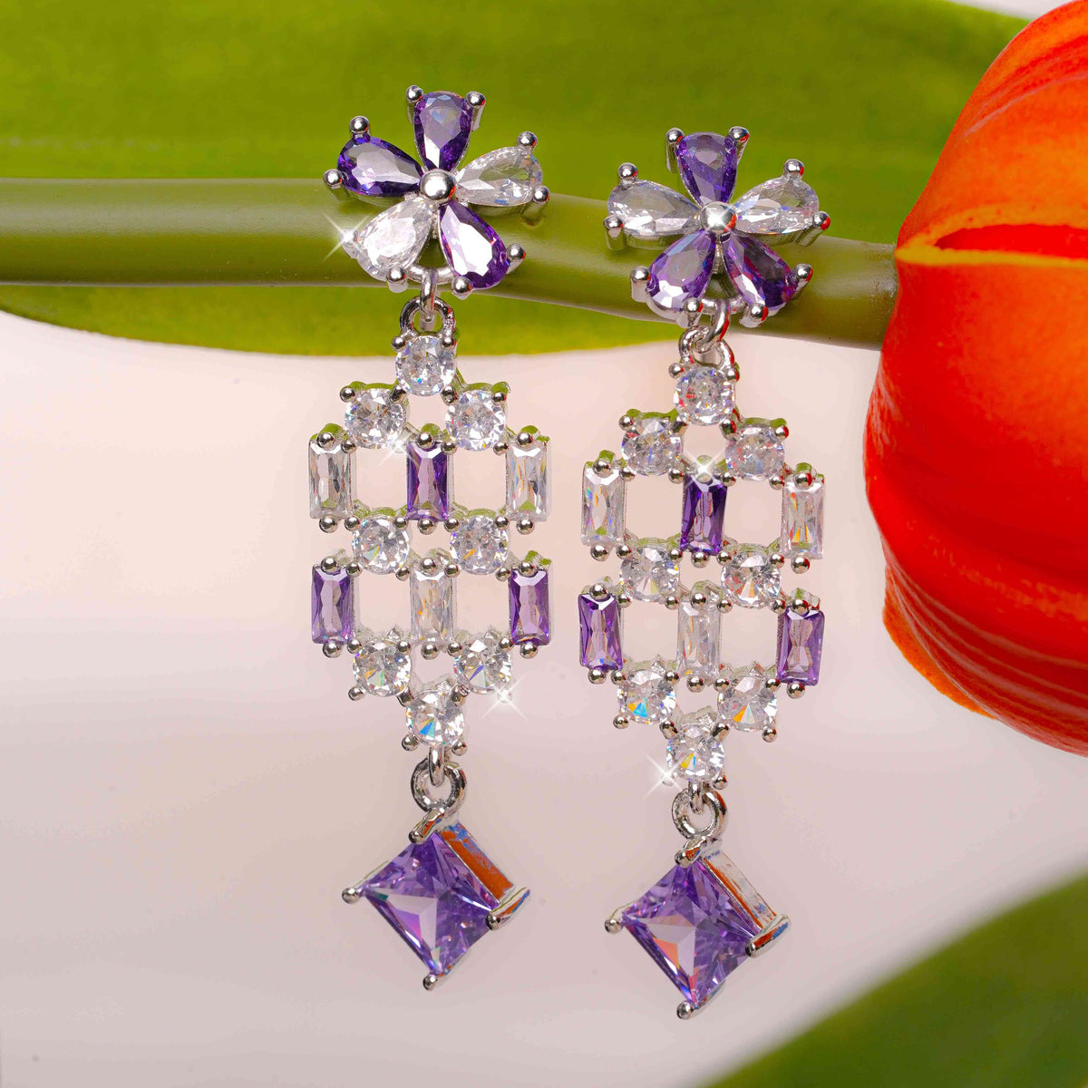 Pair of purple and clear gemstone earrings on a light background with a red flower.