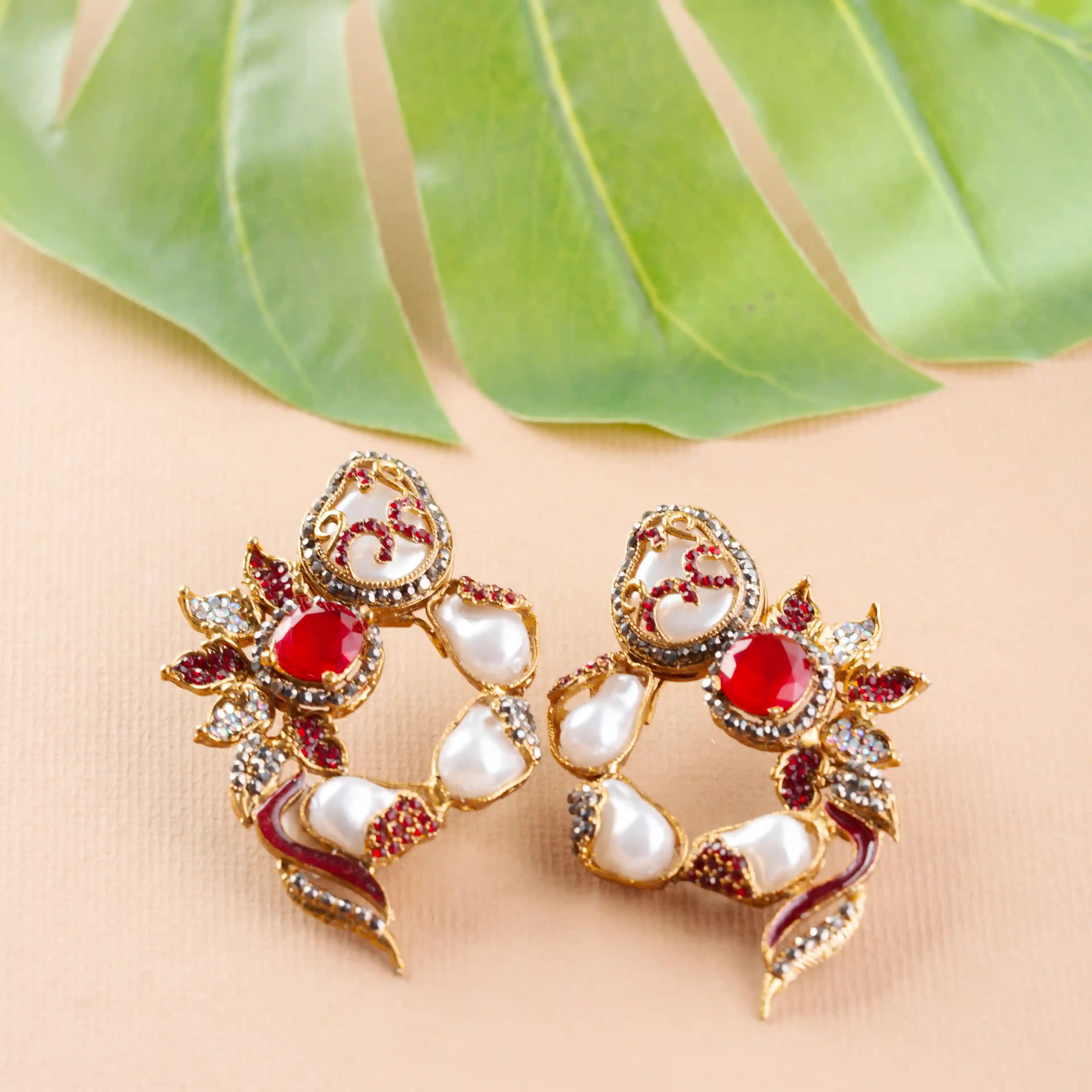 Pair of ornate earrings with red and white stones on a beige background with green leaves.