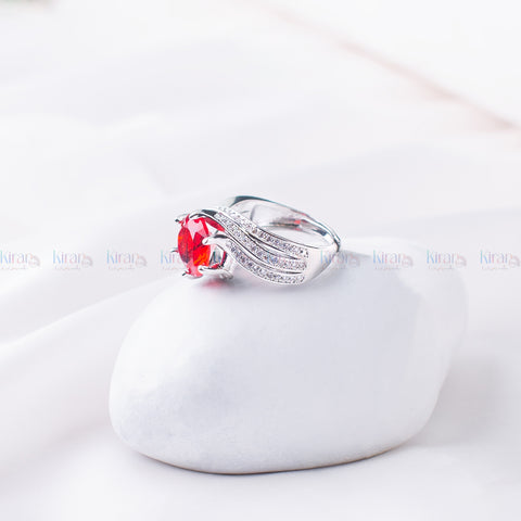An elegant silver ring featuring a red gemstone centerpiece with a halo of smaller clear stones, set on a soft white cushion.
