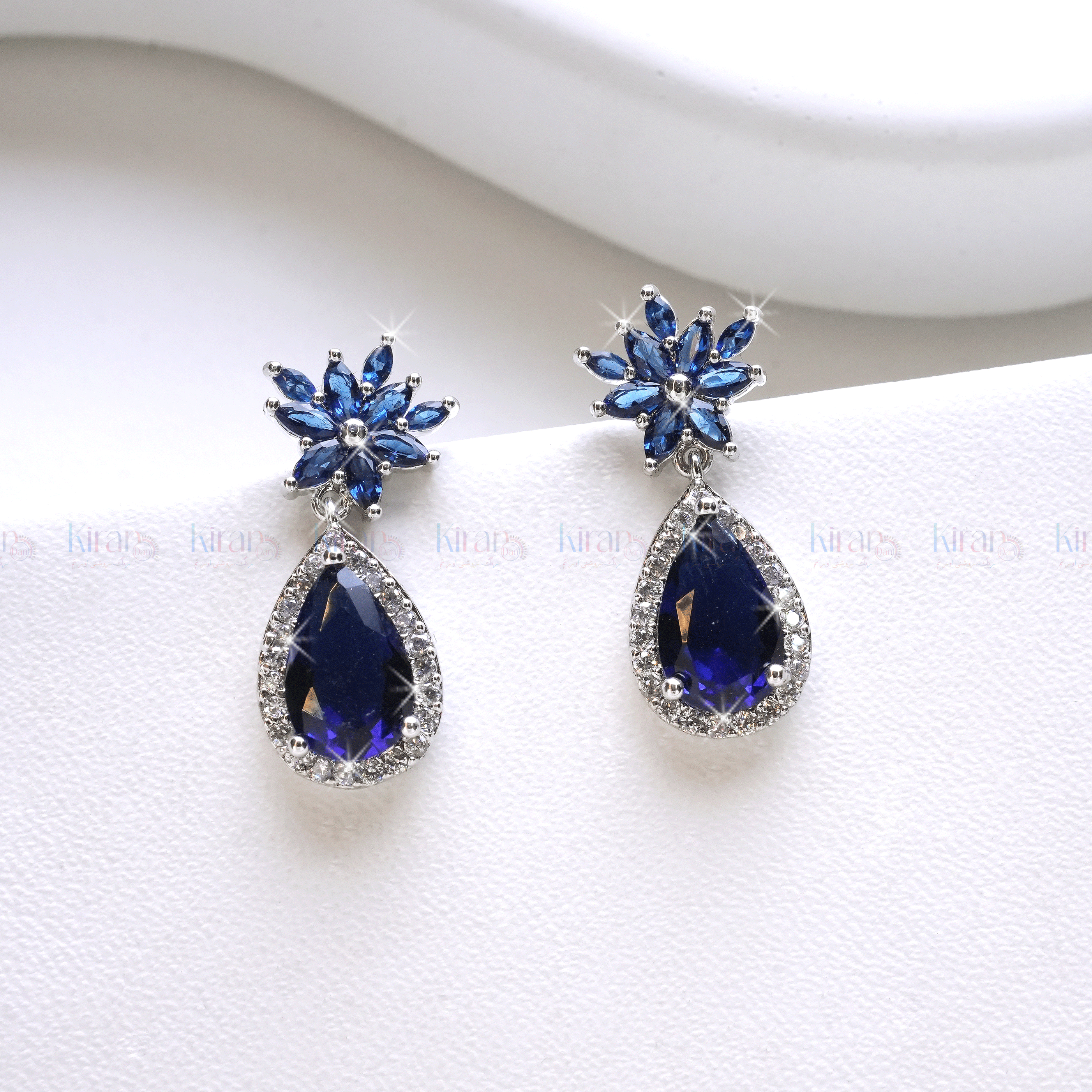 A pair of blue teardrop-shaped earrings with a floral marquise stud design and encrusted with small crystals, displayed against a white background.