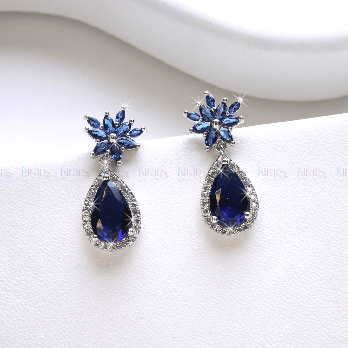 A pair of blue teardrop-shaped earrings with a floral marquise stud design and encrusted with small crystals, displayed against a white background.